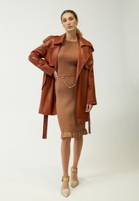 Brauner Ledermantel über einem gestrickten, gerippten Bronze-Kleid mit einem Rüschensaum. Goldene Kettendetails an der Taille. Glitzernde beigere Absätze.