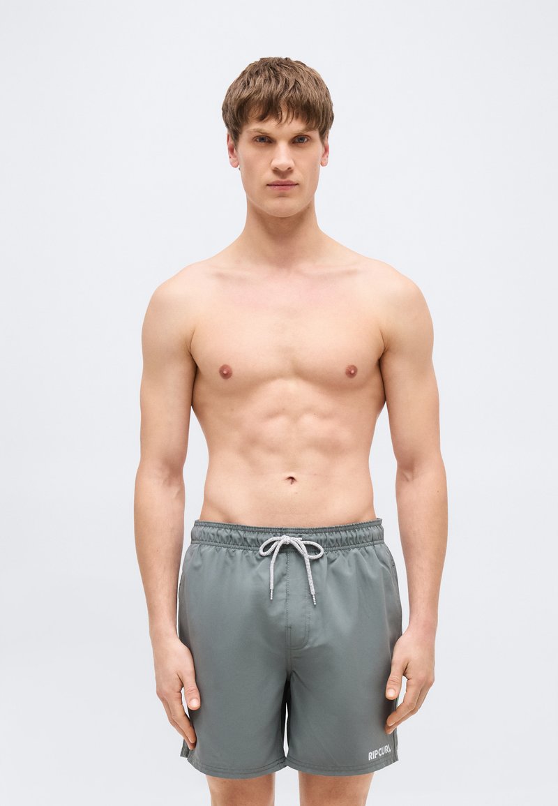 Νεαρός άνδρας που στέκεται όρθιος με το πρόσωπο προς τα εμπρός, με γυμνό στήθος, φορώντας γκρι μαγιό με λευκό κορδόνι και λογότυπο "RIP CURL" στο αριστερό πόδι.