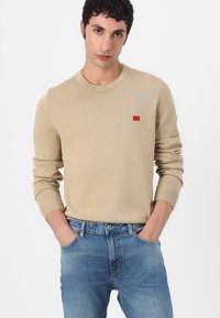Beige neulepaita, jossa on ribbikuvioiset hihansuut ja kaula-aukko, ja siinä on pieni punainen logopatch rinnan kohdalla. Yhdistetty vaaleansinisiin farkkuihin.