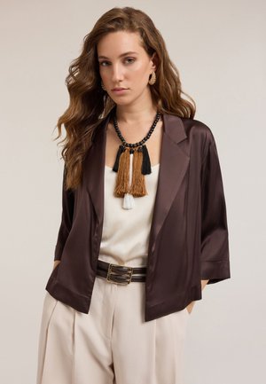 Satin brown oversized jacket with wide lapels, worn over a cream blouse. Accessories include a multi-colored tassel necklace and a wide belt.