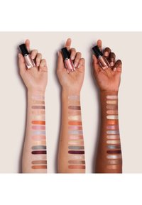 Three arms with varying skin tones hold lipstick tubes, showing matching eyeshadow swatches in multiple colors from wrist to elbow.