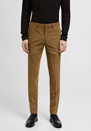 Pantalon marron ajusté porté avec des mocassins noirs et un haut noir à manches longues, debout contre un fond clair uni.