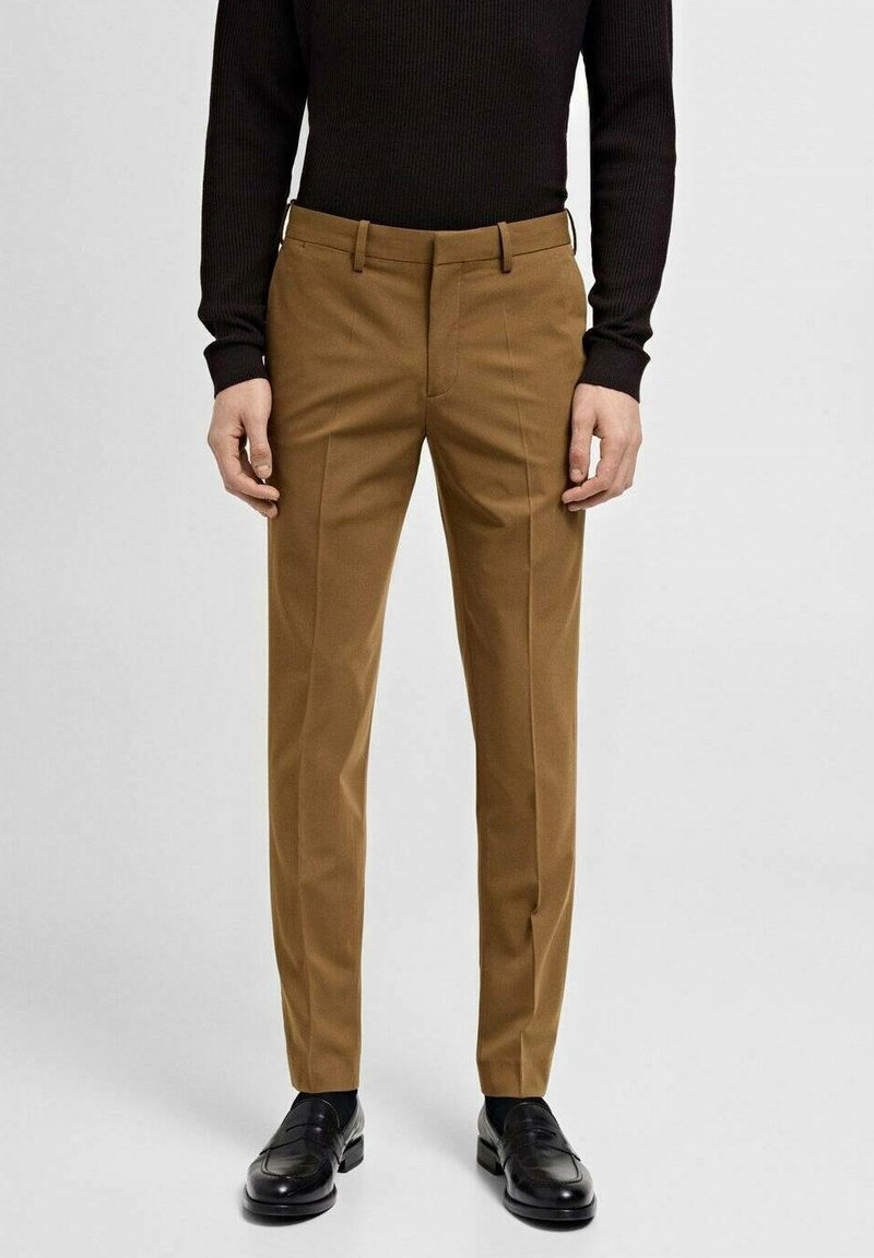 Pantalon marron ajusté porté avec des mocassins noirs et un haut noir à manches longues, debout contre un fond clair uni.