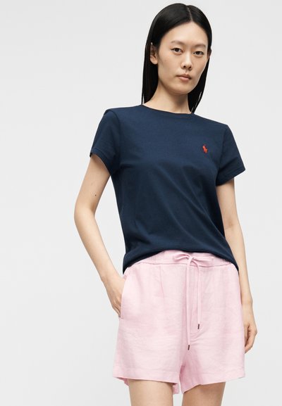Γυναίκα που φοράει μπλε σκούρο πόλο μπλουζάκι με κόκκινο λογότυπο και ανοιχτό ροζ σορτς με κορδόνι, στέκεται με το ένα χέρι στην τσέπη.