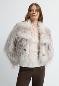Chaqueta de piel sintética en marrón claro con una textura suave, que presenta un cuello ancho y un diseño ligeramente recortado, combinada con un suéter crema y pantalones marrones.
