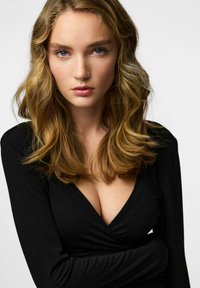 Melns apvēršanas tops ar gariem piedurknēm un dziļu V veida izgriezumu, valkāts virs brīvi krītošiem, viļņainiem plecu garuma matiem. Gaisma, neitrāla fona krāsa.