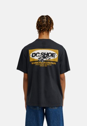 Personne avec de courtes tresses torsadées porte un t-shirt noir DC Shoes avec un motif jaune et blanc dans le dos, et un jean bleu ample.