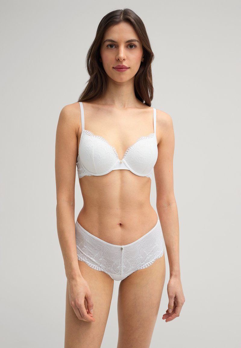 Witte kant beha met een geschulpte rand en beugels, gecombineerd met bijpassende kanten slips met een decoratieve knop in de taille.