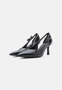 Chaussures à talons hauts en cuir verni noir avec bouts pointus et fentes. Elles comportent un petit nœud décoratif sur le côté et un talon fin.