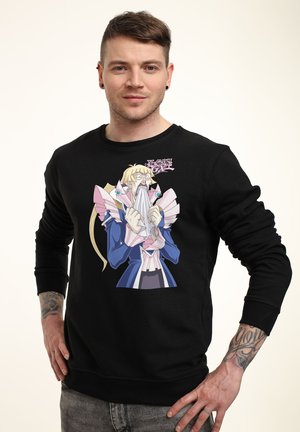 Man draagt zwarte trui met kleurrijk Yu-Gi-Oh animefiguur, houdt doek vast en staat met handen in de zij tegen een effen achtergrond.