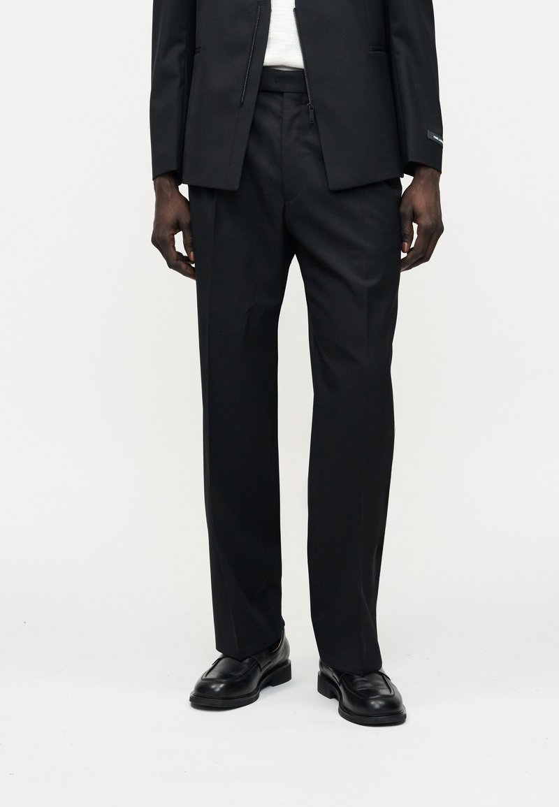 Άνδρας που φοράει μαύρο ραμμένο παντελόνι, μαύρα δερμάτινα μοκασίνια, μερικώς ορατό μαύρο σακάκι και λευκό πουκάμισο, μπροστά σε απλό φόντο.