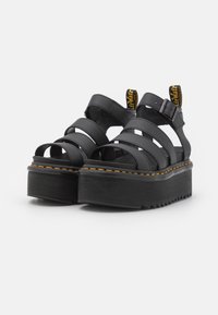 Sandales compensées noires avec larges sangles, détail de boucle et semelle en caoutchouc. Présente des coutures jaunes et une étiquette de logo au talon.