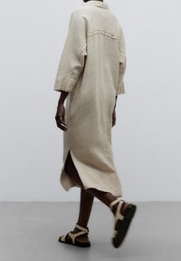 Abito in lino beige con colletto, maniche a tre quarti, spacchi laterali e vestibilità rilassata, abbinato a sandali chiari.