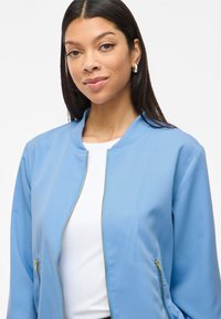 Hele sinine bomber-jakk sileda tekstuuriga, ribitud krae ja kuldsete tõmblukkudega. Omab kahte lukuga küljetaskut.