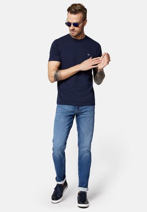 Granatowa koszulka z krótkim rękawem i okrągłym dekoltem z logo. Połączona z jasnoniebieskimi, dopasowanymi dżinsami z podwiniętymi nogawkami oraz ciemnymi trampkami.