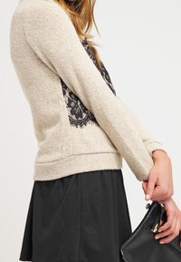 Maglione beige con tessuto strutturato, caratterizzato da un dettaglio in pizzo nero sul retro e maniche lunghe, abbinato a una gonna nera e a una pochette.