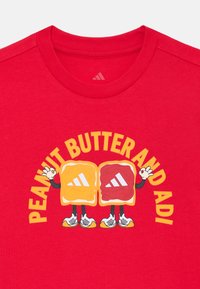 Red t-shirt featuring a graphic with two cartoon characters resembling peanut butter and jelly jars, displaying Adidas logos and text.