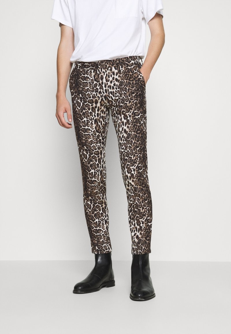 Pantalon imprimé léopard dans des tons beige et marron, coupe ajustée, tissu lisse, porté avec un t-shirt blanc uni et des bottes noires.