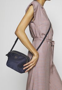 Borsa a spalla in tessuto blu navy con dettagli in oro, chiusura con zip e un logo discreto sul frontale. Texture morbida e forma rettangolare compatta.