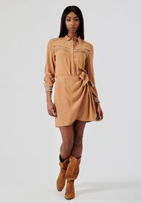 Robe boutonnière couleur tan avec une taille nouée, présentant une silhouette drapée, des manches longues, et des accents à motifs sur les poignets. Stylisée avec des bottes marron jusqu'aux genoux.