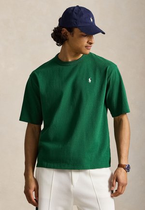 Mann trägt grünes kurzärmeliges Polohemd mit weißem Logo, weiße Hose, dunkelblaue Kappe und Armbanduhr, schaut nach unten und zur Seite.