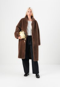 Manteau long en peluche brun avec un col châle, fermeture à boutons, porté par-dessus un haut gris clair et un pantalon noir, associé à un petit sac jaune.