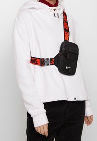 Černá taška přes rameno Nike s texturovaným povrchem, vybavená nastavitelným červeným popruhem s nápisem "NIKE", umístěná na bílé mikině s kapucí.