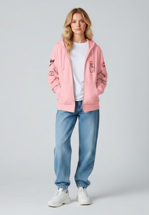 Felpa rosa con zip e stampe grafiche, abbinata a jeans larghi blu chiaro. Sneakers bianche completano l'outfit. Sfondo semplice.