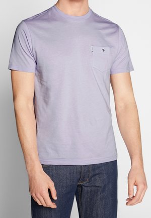 Mand iført lyse lilla kortærmet t-shirt med brystlomme og mørkeblå jeans mod en ensartet baggrund.