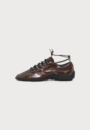 Basket en cuir brillant marron foncé avec semelle noire, laçage noir à l'avant et sangles réglables à la cheville sur fond blanc.