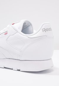 White leather sneaker with reinforced seams, padded collar, textured finish, and grey Reebok logo on the heel. Minimalist design and low-profile sole.