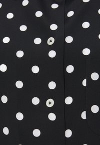 Black shirt with white polka dots, featuring a button closure and a breast pocket. Smooth fabric with a subtle sheen.