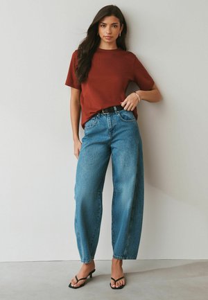 Frau trägt ein rostfarbenes T-Shirt, das in hoch taillierte, lockere blaue Jeans gesteckt ist, dazu ein schwarzer Gürtel und schwarze Sandalen mit Absatz, steht vor einer weißen Wand.