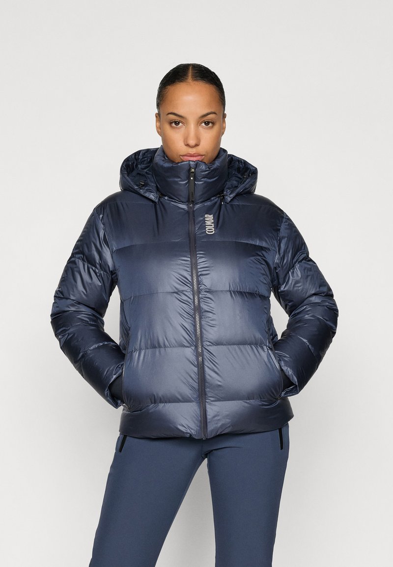 Navy puffer jacket with a high collar, front zip, and hood. Features quilted pattern and a smooth, shiny texture. Logo on the chest.