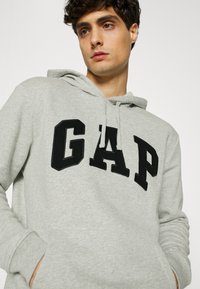 Gray hoodie featuring a kangaroo pocket, drawstring hood, and large black "GAP" logo. Made of soft cotton blend with a textured finish.