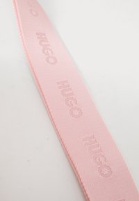 Ružová tkaná stuha so zlatočerveným embosovaným logom "HUGO" s textúrovaným povrchom a čistými hranami. Vhodná na doplnky a dekoratívne účely.