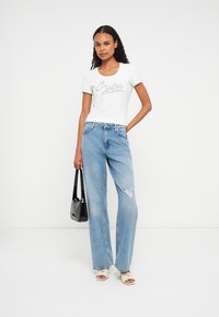 Frau trägt ein weißes T-Shirt mit Guess-Logo, hellblaue zerrissene Jeans, weiße Absatzsandalen, hält eine schwarze Handtasche und steht vor einem weißen Hintergrund.