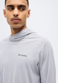 Νεαρός άνδρας που φοράει ένα ανοιχτογκρι φούτερ Columbia, κοιτάζοντας προς τα αριστερά του, μπροστά σε απλό λευκό φόντο.