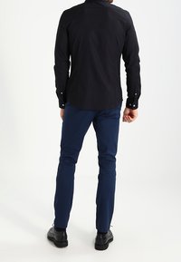 Maglietta nera a maniche lunghe con orlo arrotondato, indossata con pantaloni dritti blu navy e scarpe nere. Design semplice, senza motivi o accenti visibili.