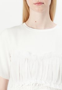 Chemise blanche avec un col rond, des manches courtes et un détail de volant froncé le long du buste qui ajoute de la texture.