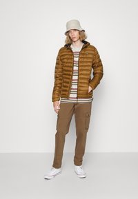 Jachetă pufosă maro cu glugă, cămașă dungată bej și gri, pantaloni cargo maro și adidași albi, asortați cu o pălărie tip bucket de culoare gri deschis.
