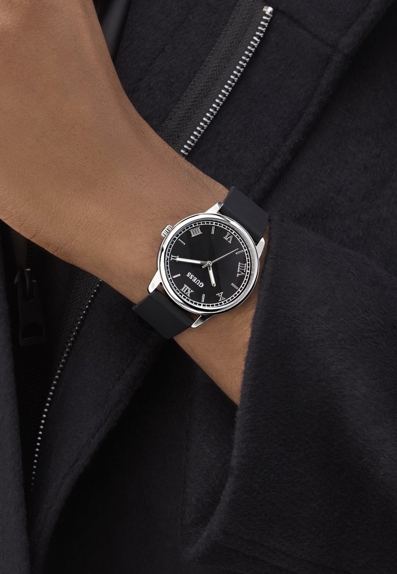 Czarny zegarek z srebrną stalową kopertą, znakiem w postaci rzymskich cyfr i czarnym gumowym paskiem. Noszony na ręce na tle ciemnej tkaniny.