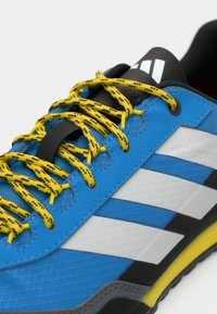 Blauer Sportschuh mit gelben und schwarzen gemusterten Schnürsenkeln, weißen Streifen an der Seite, strukturiertem Material und grauem Sohlenakzent.