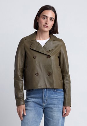 Frau trägt eine kurze olivgrüne Leder-Doppelreiherjacke über einem weißen Hemd und blauen Jeans vor einem schlichten Hintergrund.