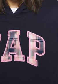 Námořnická modrá mikina s kapucí s velkým logem "GAP"; písmena mají kostkovaný vzor v růžové, námořnické a krémové barvě; růžové prošívání obkresluje písmena.