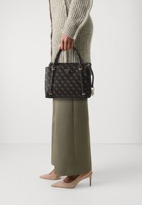 Borsa nera con motivo logo dorato, tessuto testurizzato, due manici e tracolla removibile. Abbinata a una gonna lunga oliva e tacchi beige.