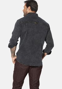 Dunkelblauses Denim-Hemd mit weicher Textur, geknöpftem Kragen, hochgekrempelten Ärmeln und kontrastierenden Nähten. Darüber getragen eine burgunderfarbene Hose.