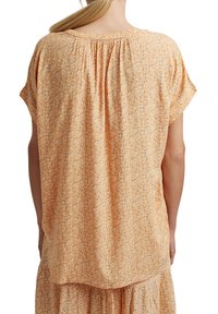 Bluse mit Blumenmuster und kurzen Ärmeln aus pfirsichfarbenem Stoff, die ein gerafftes Rückendesign aufweist. Das Material hat eine weiche Textur und eine fließende Silhouette.