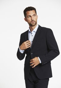 Man die een navy blazer met één knoop aanpast over een lichtblauw overhemd, terwijl hij naar de zijkant kijkt, tegen een effen witte achtergrond.