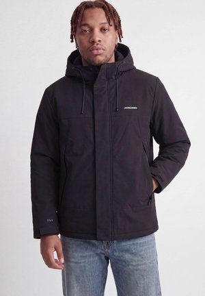 Homme aux cheveux tressés, portant une veste noire à capuche Jack & Jones et un jean bleu, debout devant un fond clair uni.
